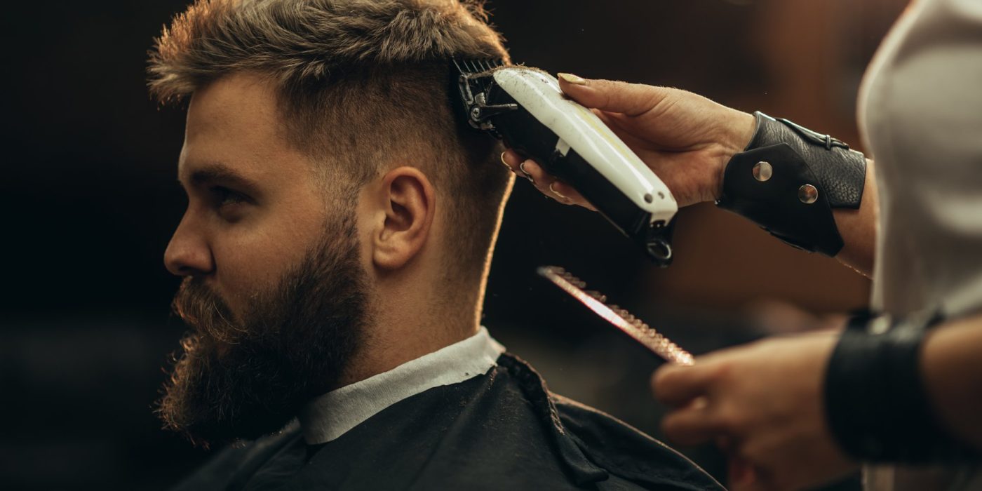 Young bearded man getting haircut by hairdresser while sitting in chair at barbershop