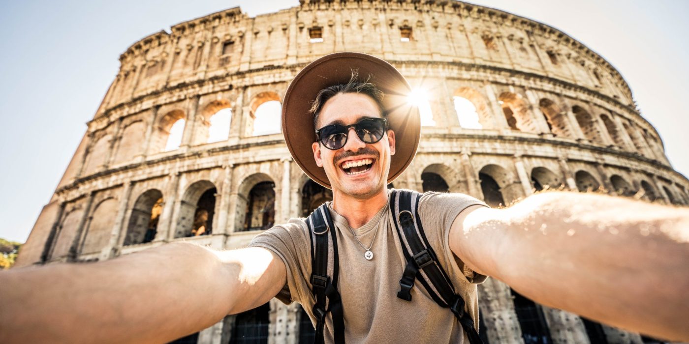 Smiling tourist visiting Coloseum in Rome, Italy - Handsome young man taking selfie with smart mobile phone device - Traveler with backpack enjoying summer vacation - Travel and technology concept